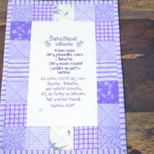 Závěsný recept na šlehačkovou bábovku je krásně vyšitý na látce s patchworkovým okrajem v odstínech fialové a bílé. Text receptu je v češtině a je vyšitý na látce. 1