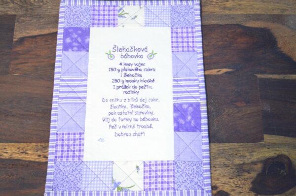 Závěsný recept na šlehačkovou bábovku je krásně vyšitý na látce s patchworkovým okrajem v odstínech fialové a bílé. Text receptu je v češtině a je vyšitý na látce. 1